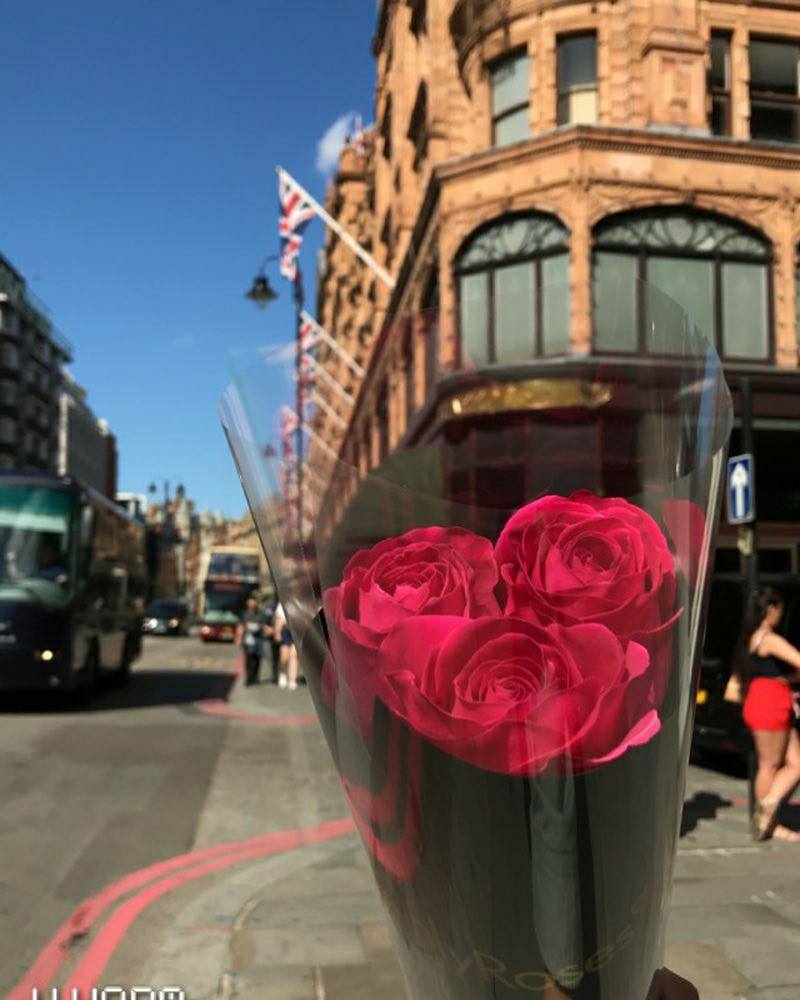 Bouquet of red roses in a transparent vase held in front of a city street with a building and flag in the background.
