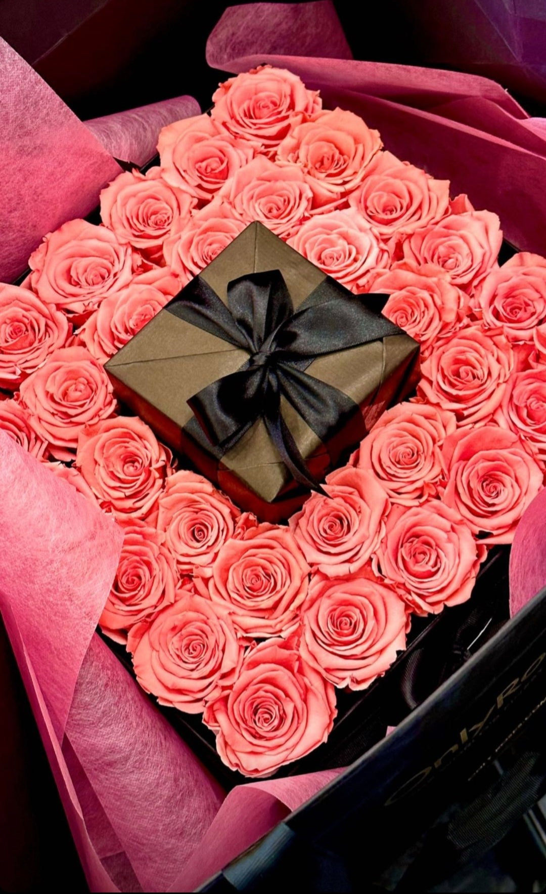 Gift box with a black ribbon surrounded by pink roses arranged in a heart shape.